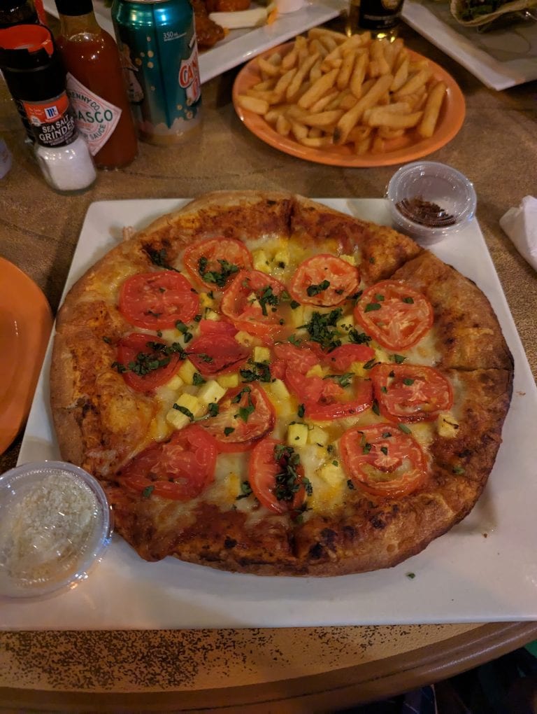 A large pepperoni and pineapple pizza is served on a white plate alongside a small cup of grated cheese and a cola in a plastic cup at a dining table in Roatan, Honduras.