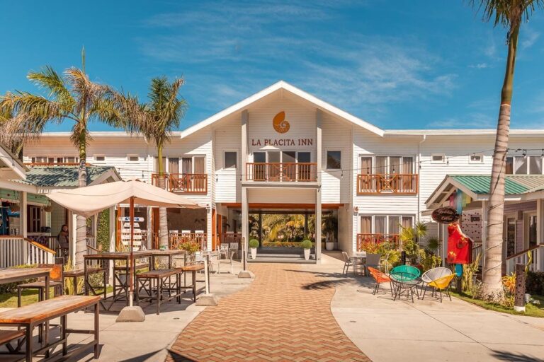 The image shows La Placita Inn, a white multi-story beachfront hotel in Roatan, Honduras, surrounded by palm trees, outdoor seating, and a clear blue sky.