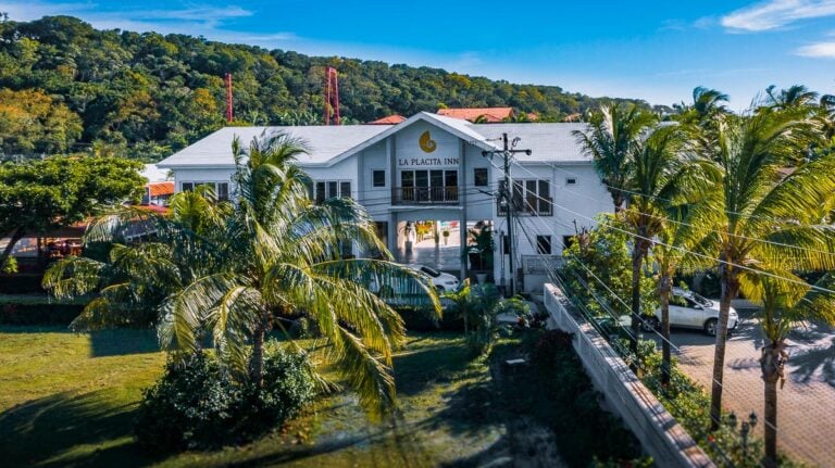 A white two-story inn named La Placita Inn is surrounded by lush palm trees and tropical greenery in Roatan, Honduras, with a parking lot and hillside covered in dense trees in the background under a bright blue sky.