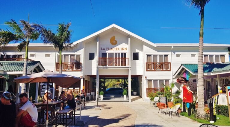 People dining and socializing at an outdoor area in front of a white building with balconies and the sign "La Placita Inn" on Roatan, Honduras, under a clear blue sky with palm trees.