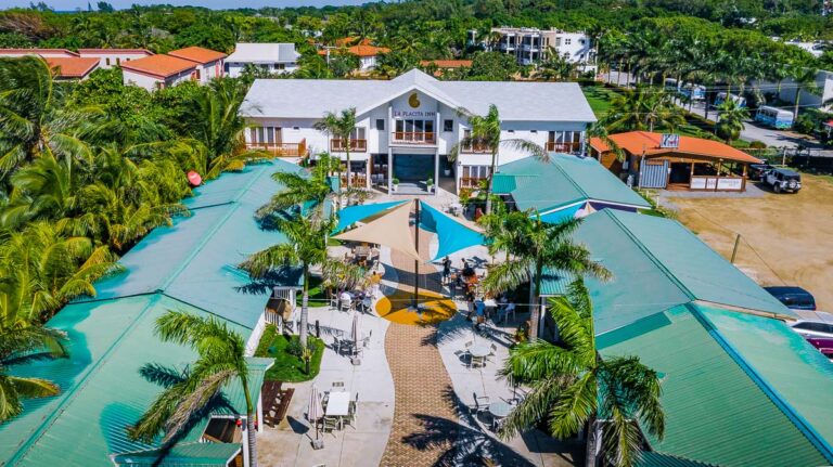 An aerial view of La Placita Inn in Roatan, Honduras, showing a courtyard with outdoor seating under colorful umbrellas, surrounded by lush palm trees and a modern white building with balconies in the background.