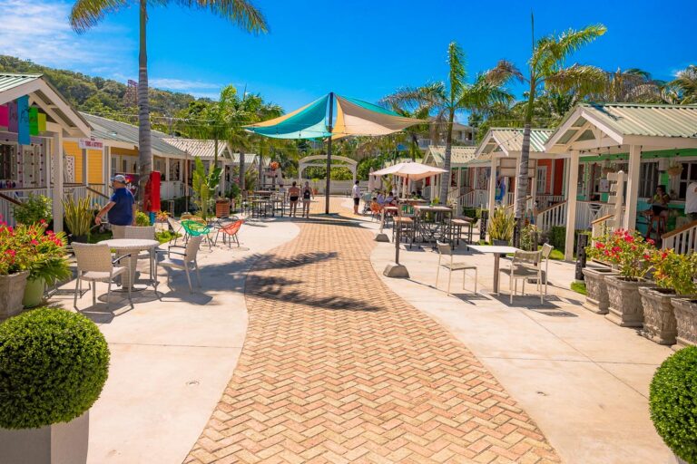 Colorful small retail shops with outdoor seating, tropical plants, and palm trees line a brick-paved walkway in a lively shopping area on Roatan, Honduras.