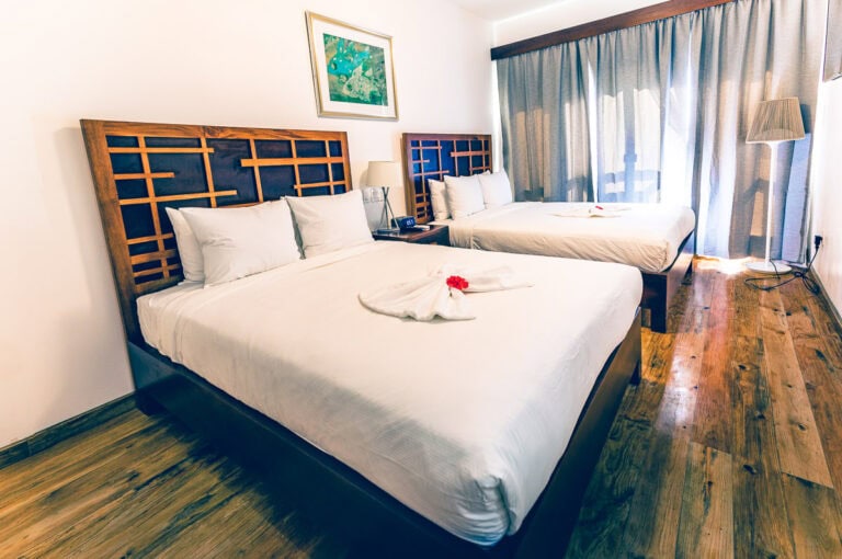 Two neatly made beds with wooden headboards are situated in a bright, modern hotel room on Roatan, Honduras, featuring white linens, decorative towel art with flowers, a framed artwork on the wall, a bedside lamp, a large window with curtains, and wooden flooring.
