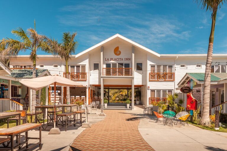 A white two-story hotel with signs reading "La Placita Inn," surrounded by palm trees, outdoor seating, colorful chairs, and umbrellas, located on a clear day in Roatan, Honduras.