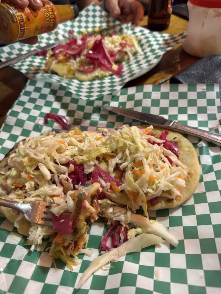 A slice of pizza topped with shredded cabbage, carrots, and red onions on a green and white checkered paper at a restaurant in Roatan, Honduras.