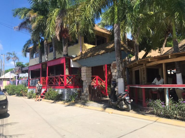 A two-story building with a yellow upper level, surrounded by tall palm trees, features a ground-floor restaurant or bar with a red wooden deck and seating area in Roatan, Honduras.
