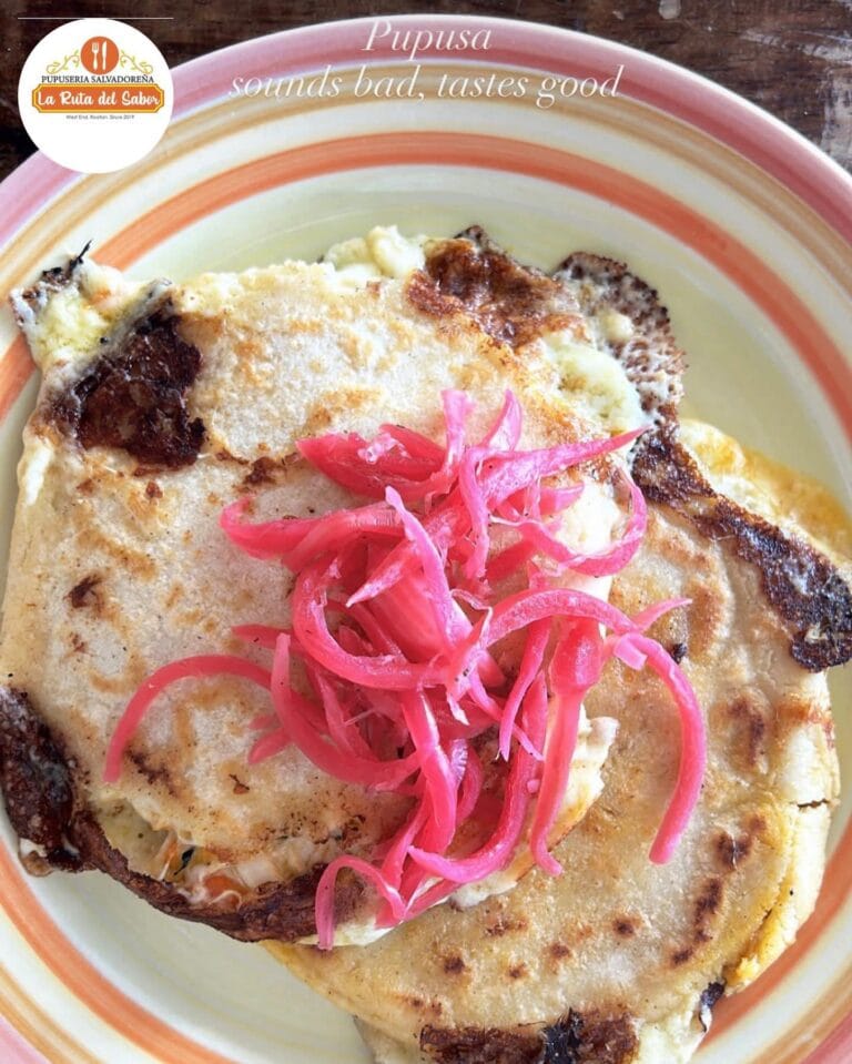 A plated Honduran dish featuring fried plantains topped with pink pickled onions, served on a colorful ceramic plate with a logo for Pupuseria Salvadoreña La Ruta del Sabor in Roatan.