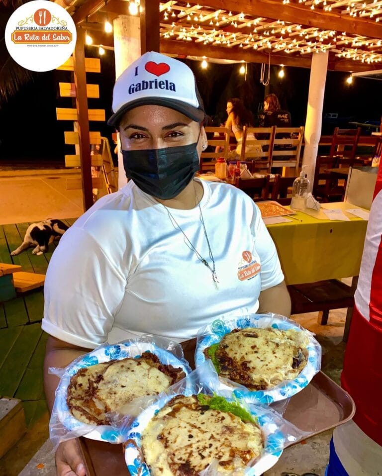 A woman wearing a "I love Gabriele" cap and face mask holds a tray of four pizza slices at a night market or outdoor restaurant in Roatan, Honduras, with string lights and wooden tables and chairs in the background.