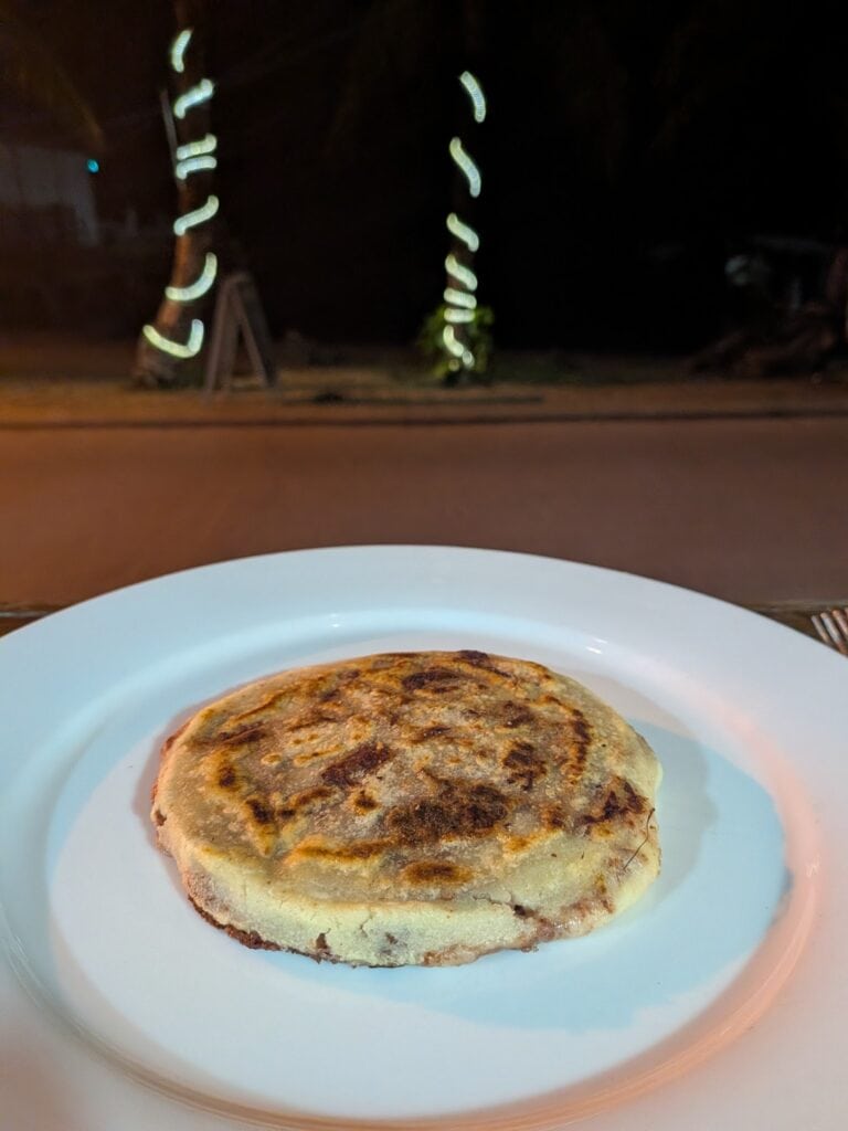 A toasted corned beef and potato patty served on a white plate with a blurred nighttime street scene in Roatan, Honduras, visible in the background.