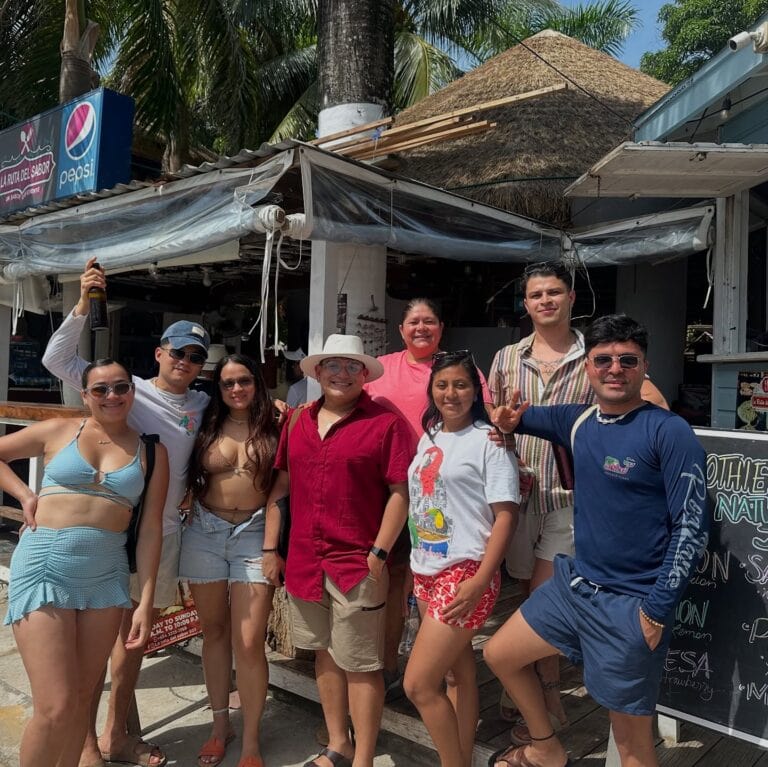 A group of nine diverse people posing together in front of a beachside restaurant or bar on Roatan in the Bay Islands of Honduras, with palm trees and a thatched roof structure visible in the background.
