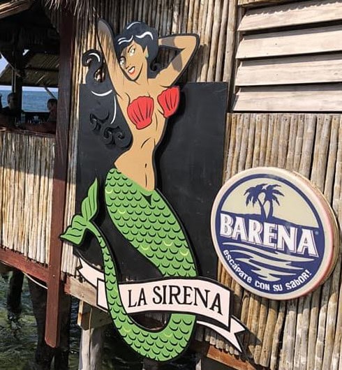 A colorful sign featuring a cartoon mermaid with long black hair, a red seashell bra, and a green fish tail, representing La Sirena at a waterfront restaurant or bar in Roatan, Honduras.