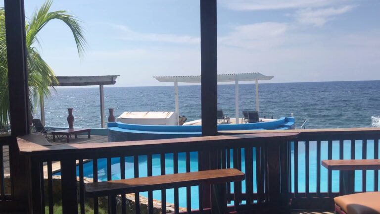 A view of the ocean from a seaside restaurant or bar in Roatan, Honduras, featuring a swimming pool, wooden lounge chairs, and a small boat with a shaded platform on the water.