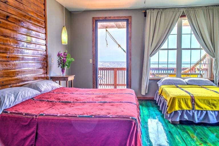 A bedroom in a Roatan, Honduras hotel or rental with two beds, one draped in a red bedspread and the other in a yellow one, overlooking the ocean through large windows and an open door leading to a balcony.