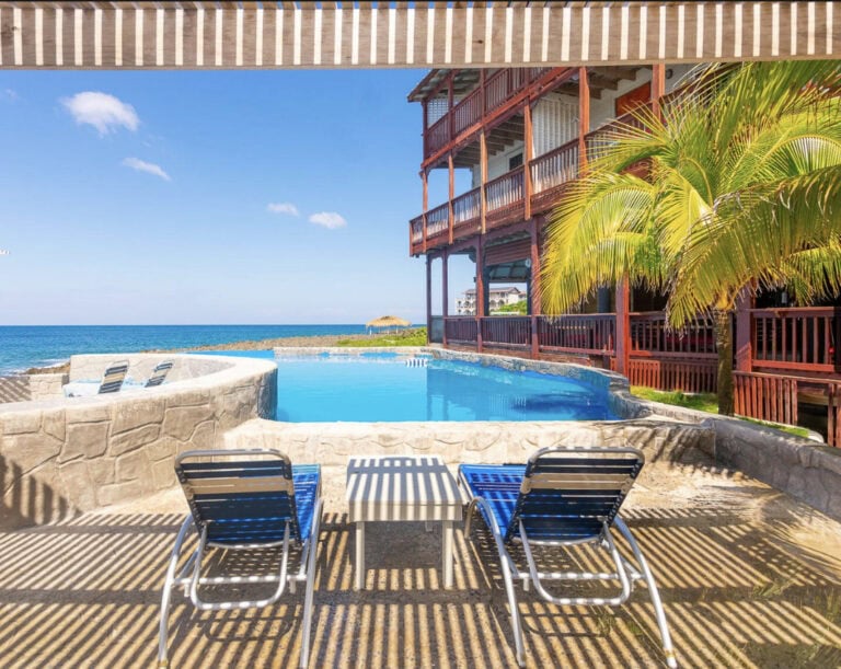 A view of a poolside area at a resort in Roatan, Honduras, featuring two blue lounge chairs and a small table on a shaded patio, with a large palm tree, a wooden building with multiple balconies, and an ocean in the background under a clear blue sky.