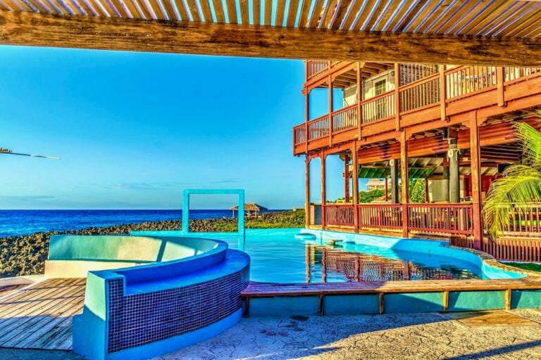A swimming pool located on the beachfront of Roatan, Honduras, with a wooden deck and patio area, overlooking the ocean with a rocky shoreline and clear blue sky.