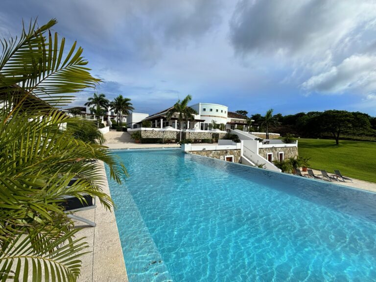 A luxurious hotel pool area with clear blue water, surrounded by white buildings, lush green palm trees, and shaded lounge chairs, set against a backdrop of partly cloudy skies in Roatan, Honduras.