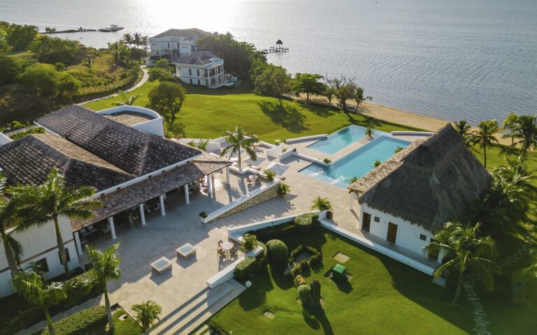 Aerial view of a luxurious beachfront resort in Roatan, Honduras, featuring multiple white buildings with thatched and tiled roofs, a large infinity swimming pool, lush green lawns, palm trees, and a sandy beach overlooking the Caribbean Sea.