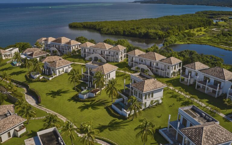 White multi-story villas with terracotta tiled roofs are surrounded by lush green lawns and palm trees on a coastal property in Roatan, Honduras, overlooking the ocean and nearby islands.