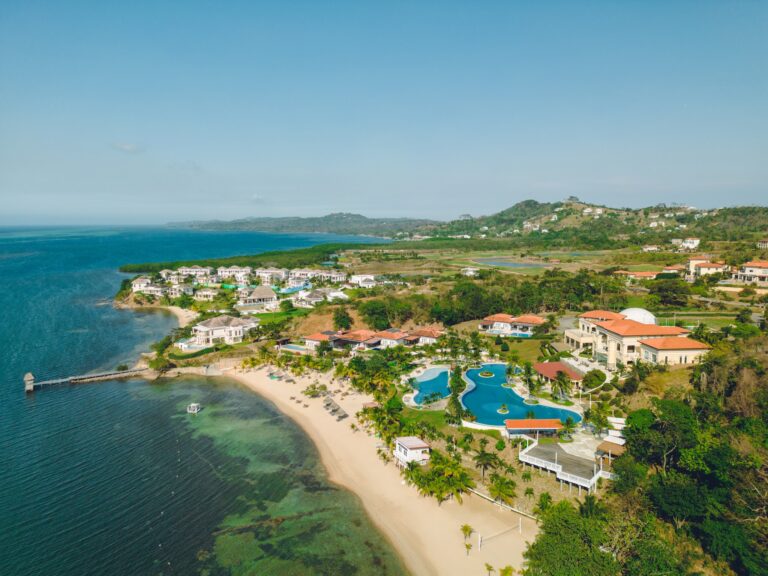 Aerial view of a beachfront resort with white villas, swimming pools, and lush greenery along the coast of Roatan in Honduras, with hills and residential areas in the background.
