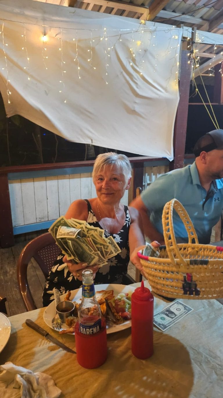A smiling woman at a dining table in Roatan, Honduras, holding a large stack of dollar bills with a plate of food, condiments, a bottle of soda, and a basket on the table, with string lights and a wooden-partitioned background.