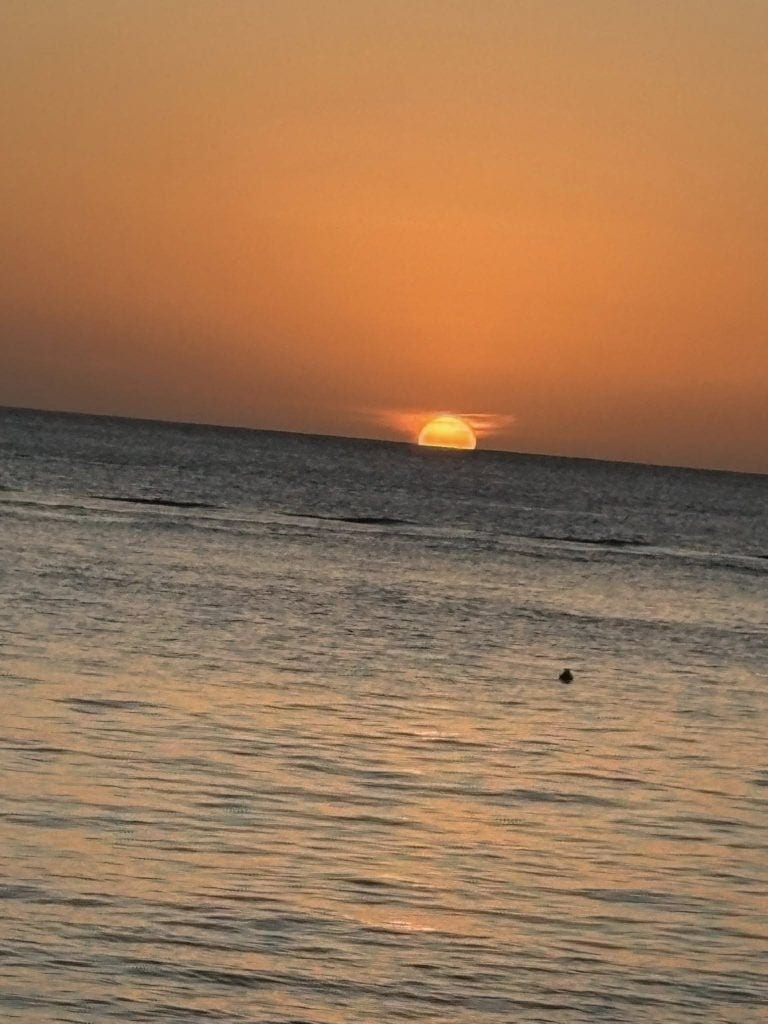 A sunset over the ocean with the sun partially below the horizon and the sky painted in warm orange tones, near a coastline in Roatan, Honduras.