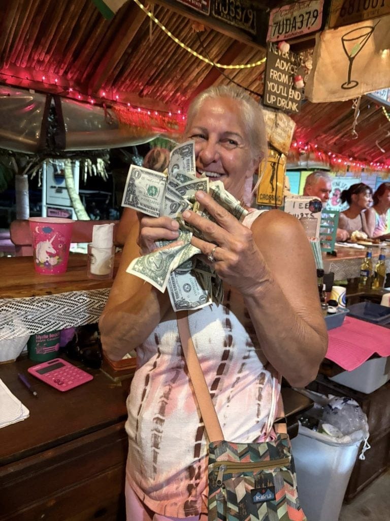 A smiling elderly woman holding U.S. dollar bills sits at a wooden table inside a lively bar or restaurant in Roatan, Honduras, decorated with colorful lights, signs, and a chalkboard with humorous messages.
