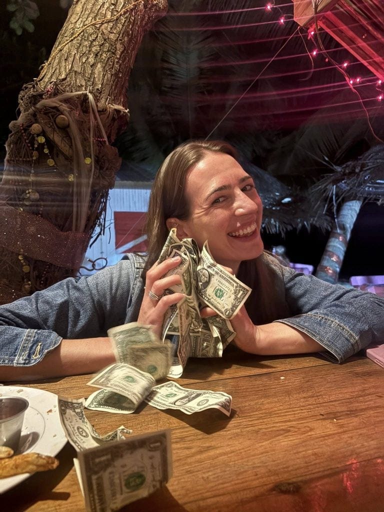 A smiling woman in a denim jacket holds dollar bills while sitting at a wooden table in a dimly lit restaurant or bar in Roatan, Honduras, with crumpled cash and a plate of food in front of her and colorful string lights hanging overhead.