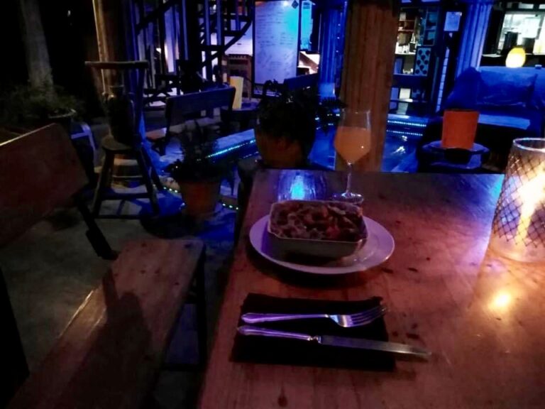 A cozy outdoor dining area in Roatan, Honduras, features a wooden table set with a bowl of food, a glass of drink, cutlery, and candlelight, with a background of potted plants, chairs, and indoor decor visible through large glass doors.