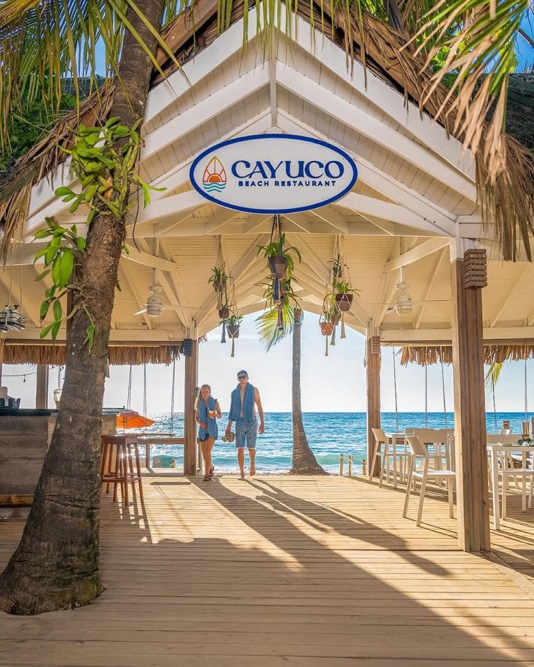 A beachfront open-air restaurant with a thatched roof, hanging plants, and white furniture overlooking the Caribbean Sea in Roatan, Honduras.