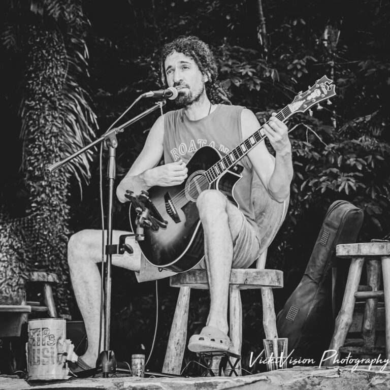 A man with long, curly hair plays an acoustic guitar and sings into a microphone during a live outdoor performance in a lush, tropical setting on Roatan, Honduras.