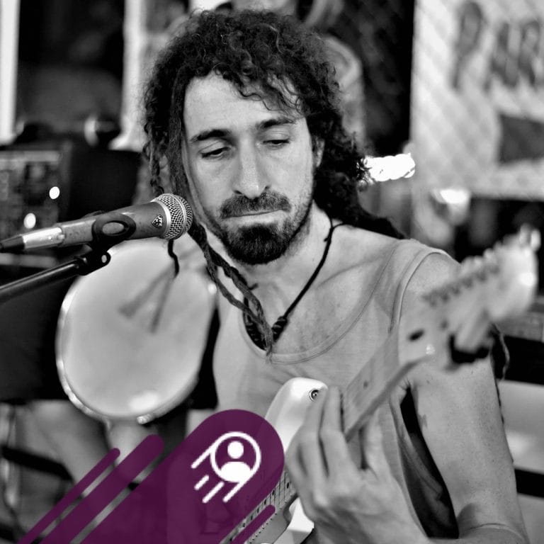 A man with curly hair and a beard is playing an acoustic guitar and singing into a microphone in a casual indoor setting on Roatan, Honduras, with blurred background elements.