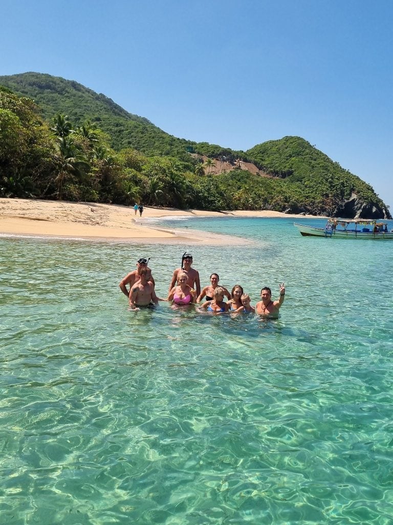 A group of people swimming and enjoying the clear turquoise waters near a sandy beach with lush green hills in Roatan, Honduras, visible in the background.