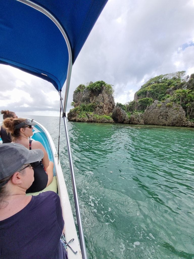 People are seated on a boat with a blue canopy, enjoying a scenic view of rocky, lush green islands and cloudy skies in the waters near Roatan, Honduras.