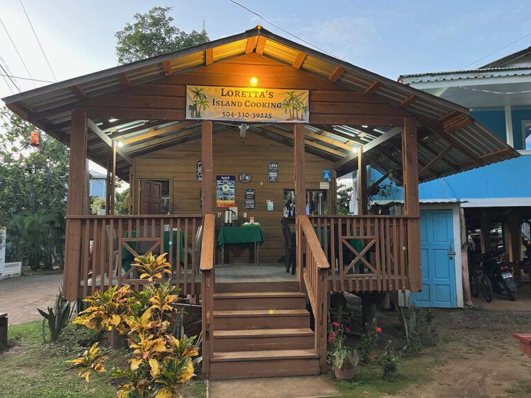 A small, elevated wooden restaurant with a sign reading "Loretta's Island Cooking" is set against a tropical background in Roatan, Honduras, featuring a front porch with steps, outdoor seating, and decorative plants, illuminated by warm lighting.