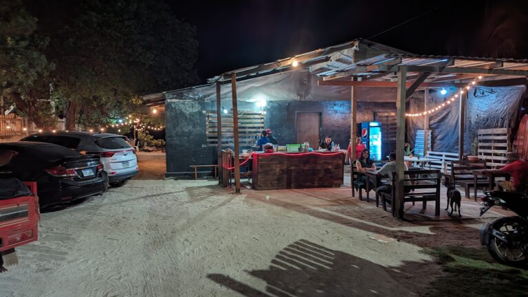 A nighttime view of an open-air restaurant or bar in Roatan, Honduras, featuring a wooden structure with string lights, surrounded by parked cars, with people dining and a Pepsi vending machine visible in the background.