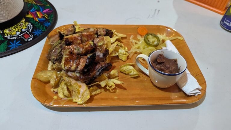 A plate of grilled pork ribs with fried plantains, pickled vegetables, and a small cup of refried beans set on an orange tray at a restaurant in Roatan, Honduras.