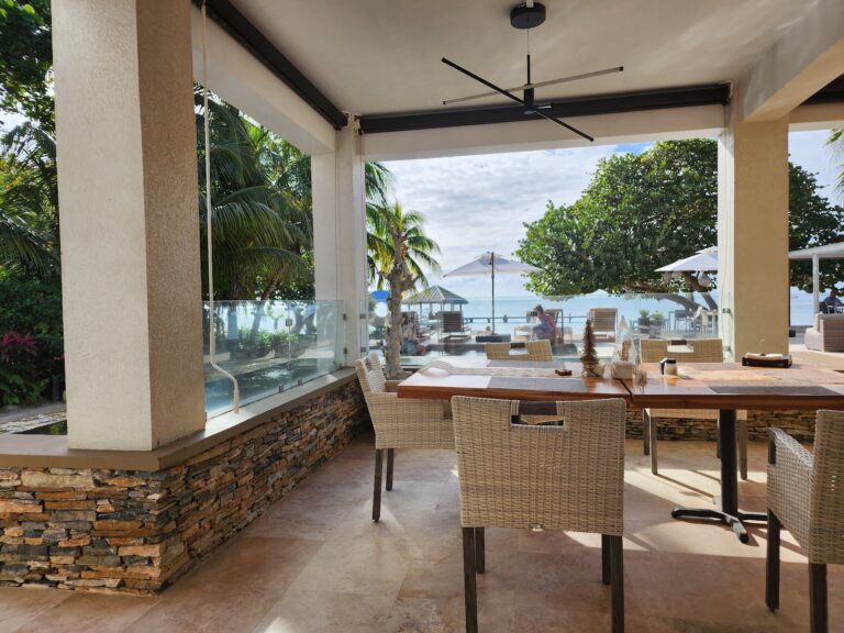 A shaded outdoor dining area in Roatan, Honduras, features wicker chairs, a wooden table, and a view of palm trees, umbrellas, and the ocean in the background.