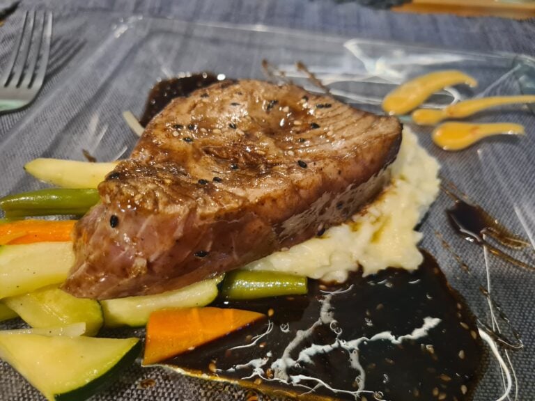 A grilled steak served with mashed potatoes and colorful roasted vegetables on a black plate, set on a dark table in Roatan, Honduras.