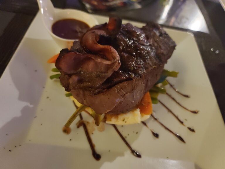A plated serving of grilled steak topped with sautéed mushrooms and accompanied by a side of vegetables and sauce on a white dish in Roatan, Honduras.
