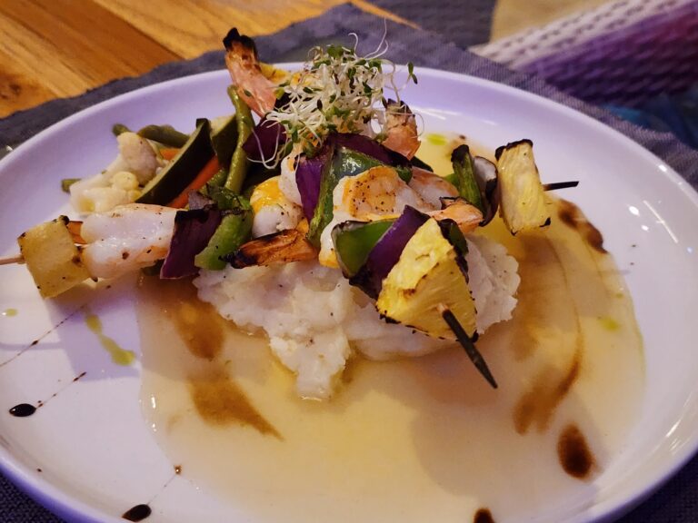 A skewered seafood and vegetable dish garnished with microgreens is served on a white plate, with Roatan, Honduras, visible in the background.