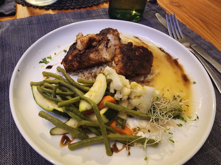 A plate of seasoned fried fish served over rice with cooked green beans, sliced zucchini, shredded cabbage, microgreens, and a drizzle of dark sauce, set on a dark placemat on a wooden table in Roatan, Honduras.