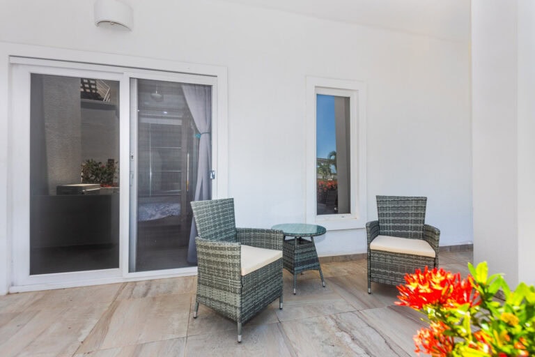 A white-walled outdoor patio in Roatan, Honduras, features two woven rattan armchairs with beige cushions and a small round glass-top table, set against a sliding glass door and a narrow window, with vibrant red flowers in the foreground and palm trees visible outside.