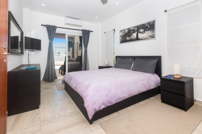 Modern bedroom in a Roatan guesthouse featuring a bed with purple bedding, black furniture, and wall art depicting a tree silhouette in Honduras.