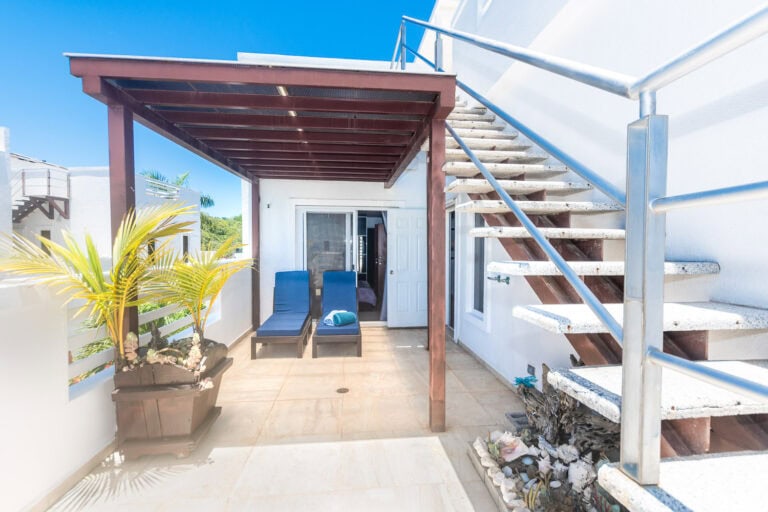 Balcony area on a tropical island in Roatan, Honduras featuring a white exterior wall, two blue lounge chairs with towels, potted tropical plants, and an outdoor staircase leading to the upper level under a wooden pergola with clear blue sky in the background.