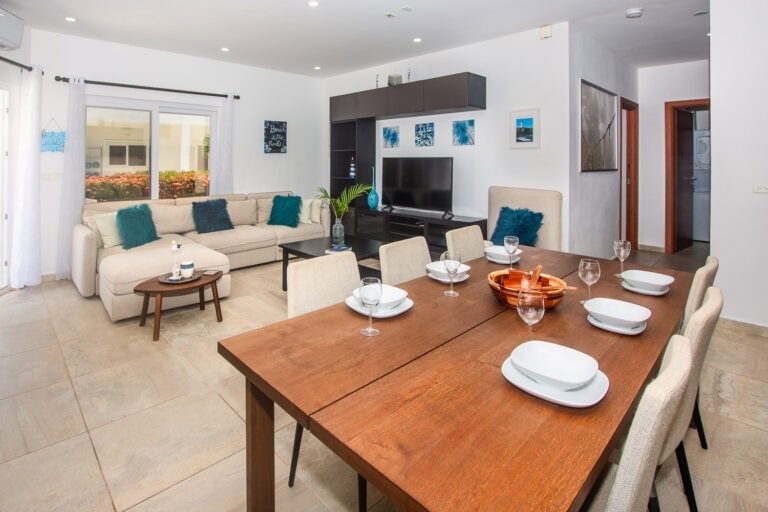 Interior view of a living and dining area in a vacation rental in Roatan, Honduras, featuring a beige sofa with teal pillows, a wooden dining table set for six, and a flat-screen TV with beach-themed artwork on the white walls.