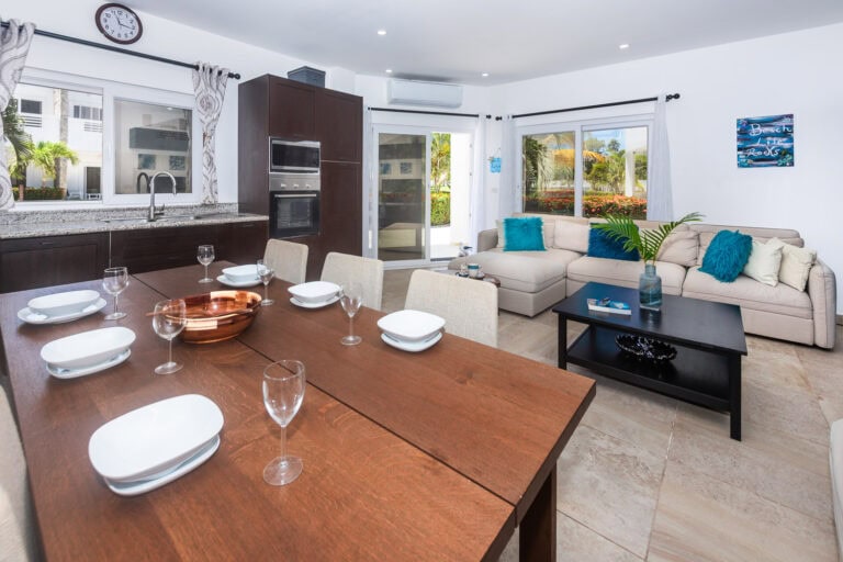 Interior view of a modern living and dining area in a house on Roatan, Honduras, featuring a kitchen with dark wood cabinets, a dining table set for six, a beige sectional sofa with colorful throw pillows, and large windows overlooking tropical greenery.