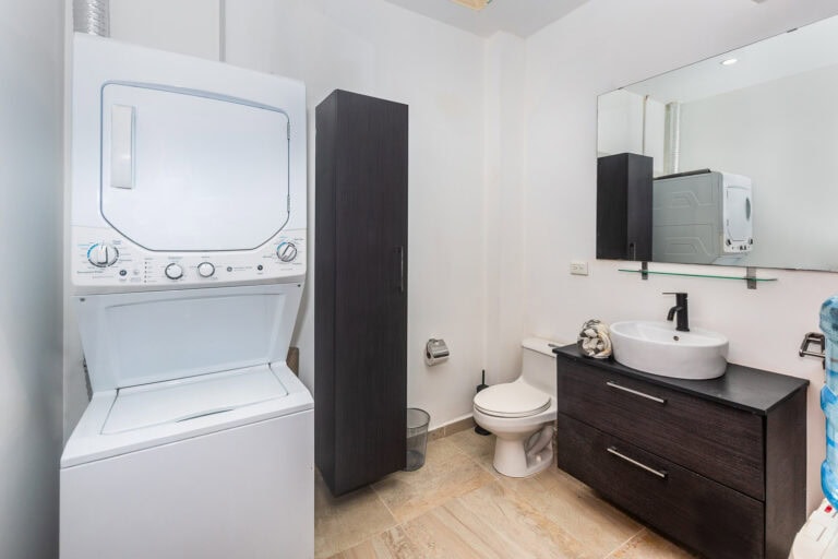 A compact bathroom in Roatan, Honduras, featuring a stacked washer and dryer, a black cabinet, a toilet, and a black vanity with a round sink and mirror.