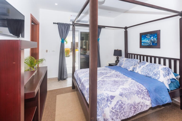 A cozy bedroom in a Roatan guesthouse with a dark wooden four-poster bed, blue and white floral bedding, black bedside lamp, and a sliding glass door leading to an outdoor patio with tropical plants in Honduras.