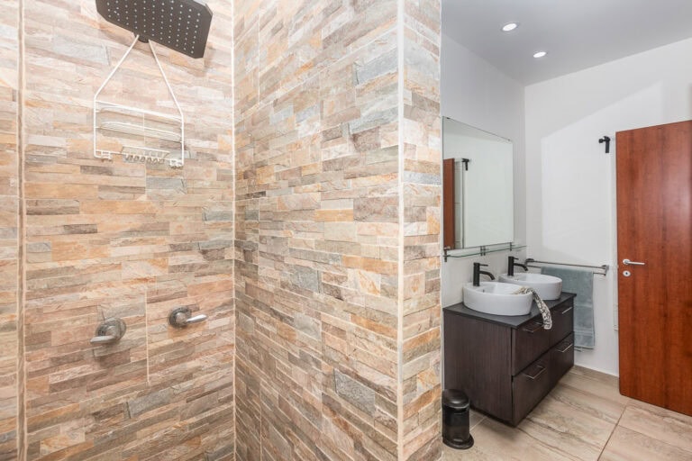 Modern bathroom with a double sink vanity, mirror, and wooden door in Roatan, Honduras, featuring a tiled shower area with beige and brown stone tiles.