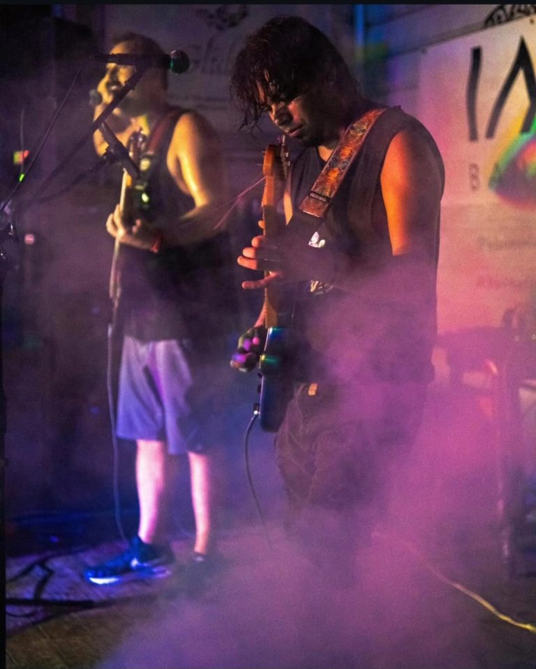 A musician with long dark hair performs on stage in Roatan, Honduras, playing an electric guitar amidst colorful lighting and haze at a live music event.
